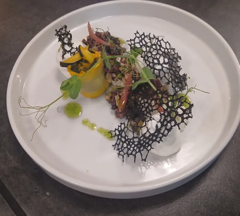 Une assiette dressée : légumes roulés, tuile noire en dentelle et herbes fraîches